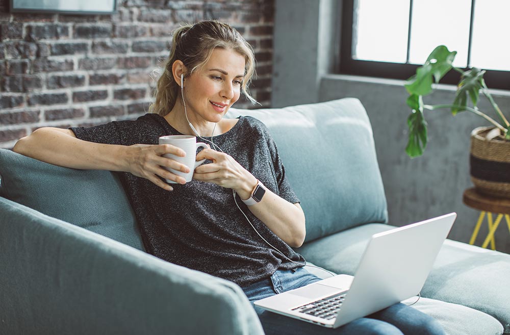 Frau sitzt auf dem Sofa mit Laptop, Kopfhörern und Tasse in der Hand